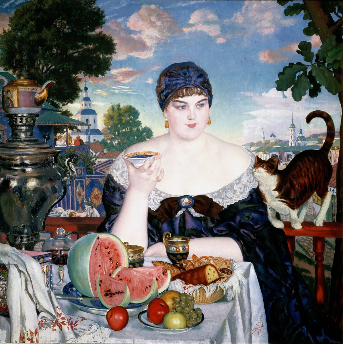 A woman takes tea on a balcony at a table laid with a brass samovar, porcelain, fruit and pastries, with town rooftops behind her.
